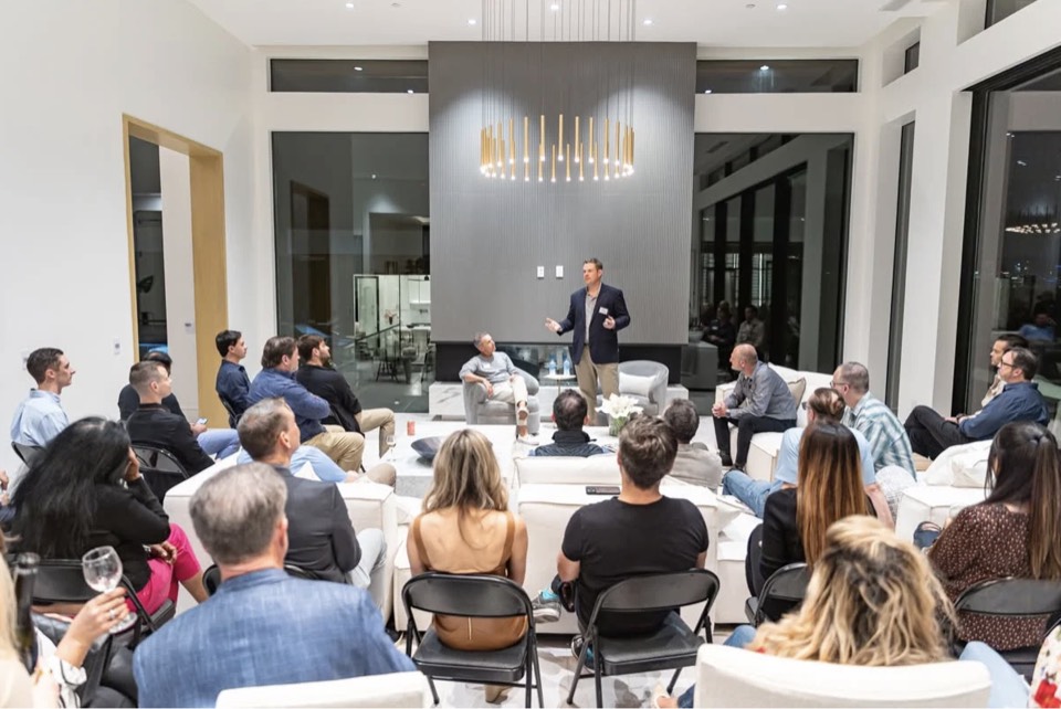 Chris Beaman standing to speak to a room of founders and investors seated on sofas and folding chairs at one of his investor dinners.