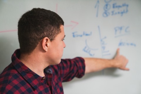 Close profile of Isaiah pointing at a whiteboard diagram of the emotional wall and prefrontal cortex, the brain model behind the dropping-guard conversation.