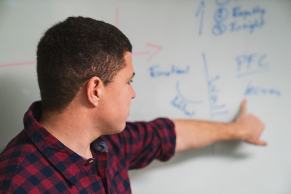 Close profile of Isaiah pointing at a whiteboard diagram of the emotional wall and prefrontal cortex, the brain model behind the dropping-guard conversation.