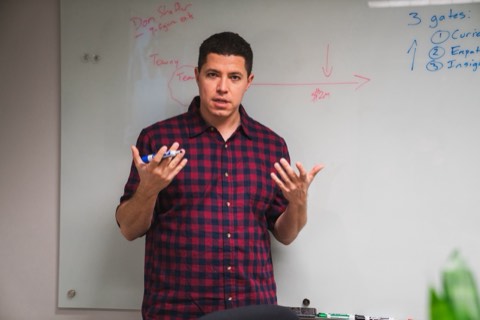 Isaiah stands at a glass whiteboard mid-explanation, both hands raised, marker in hand, with a funnel sketch in red and a numbered 3 Gates list in blue behind him.