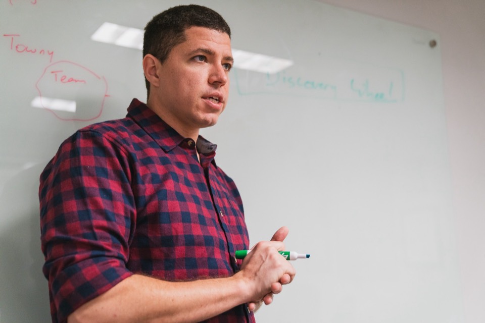 Isaiah at a whiteboard with Discovery Wheel written on it, green marker in his clasped hands, mid-sentence with a coachee off camera.
