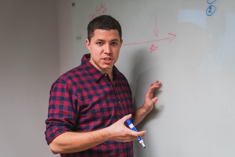 Isaiah in a three-quarter view at the whiteboard, one hand raised toward the board, talking directly to the room with a red funnel arrow beside him.