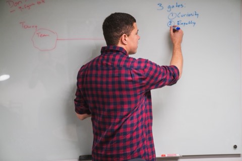 Seen from behind, Isaiah writes a numbered list on the whiteboard reading 3 gates: 1 Curiosity, 2 Empathy, in blue marker.