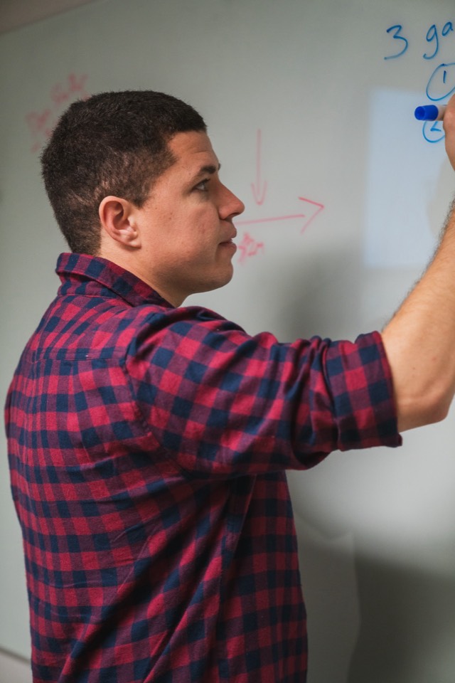 Vertical profile of Isaiah reaching up to write on the whiteboard, blue marker against the board, red funnel arrow in the background.
