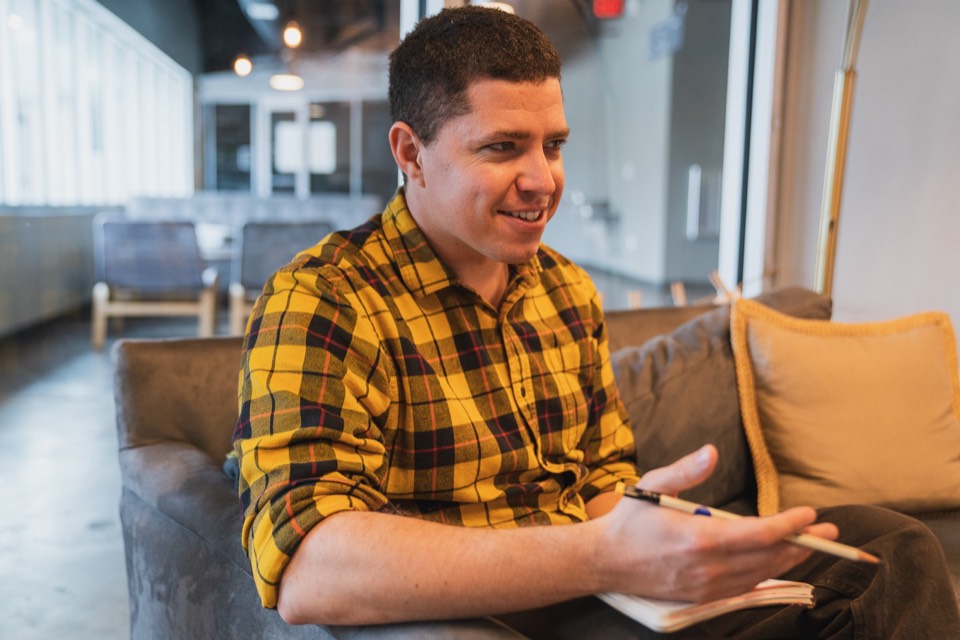 Isaiah sits on a couch with a notebook on his knee and pen in hand, smiling mid-conversation with someone off camera.