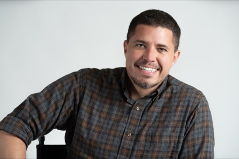 Studio headshot of Isaiah in a plaid shirt, arm resting on a chair back, smiling directly at the camera against a white background.