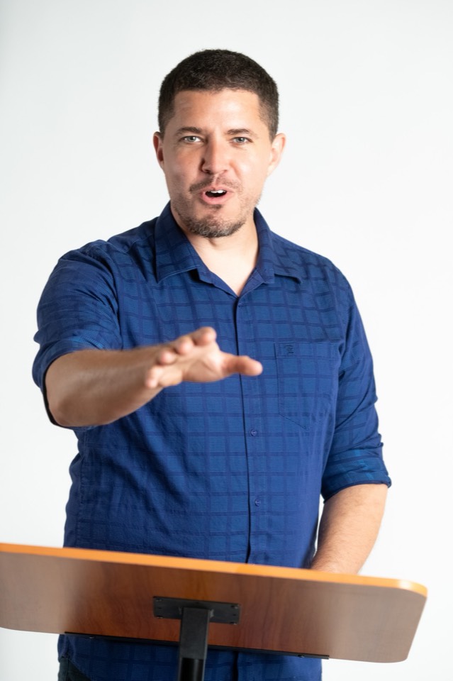 Isaiah speaks from behind a lectern, hand extended toward the audience mid-point, in a blue shirt against a white studio background.