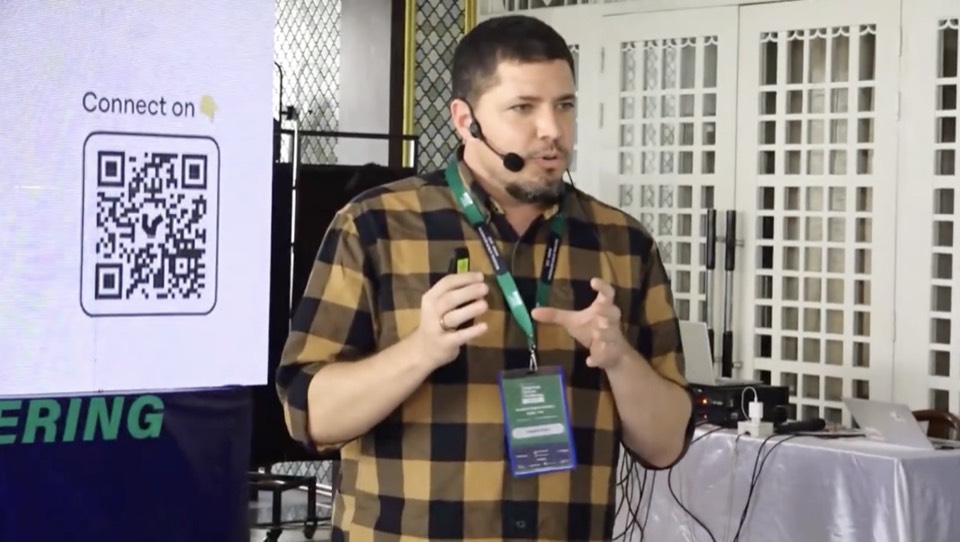 Isaiah on stage at a conference in a checked shirt and lanyard, headset mic on, both hands raised mid-gesture, a screen with a QR code behind him.